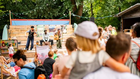 Familien mit Kindern vor einer Open-Air-Bühne,a uf der drei Musiker:innen spielen